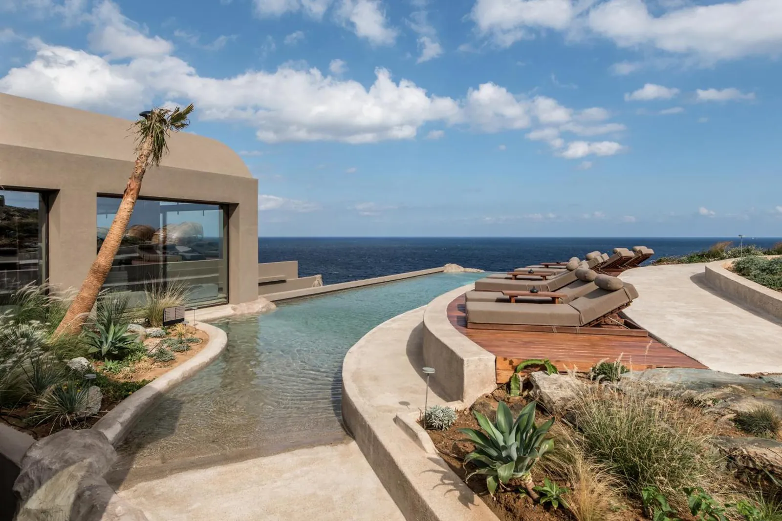 Curved infinity pool with loungers and a shaded cabana above Mononaftis Bay