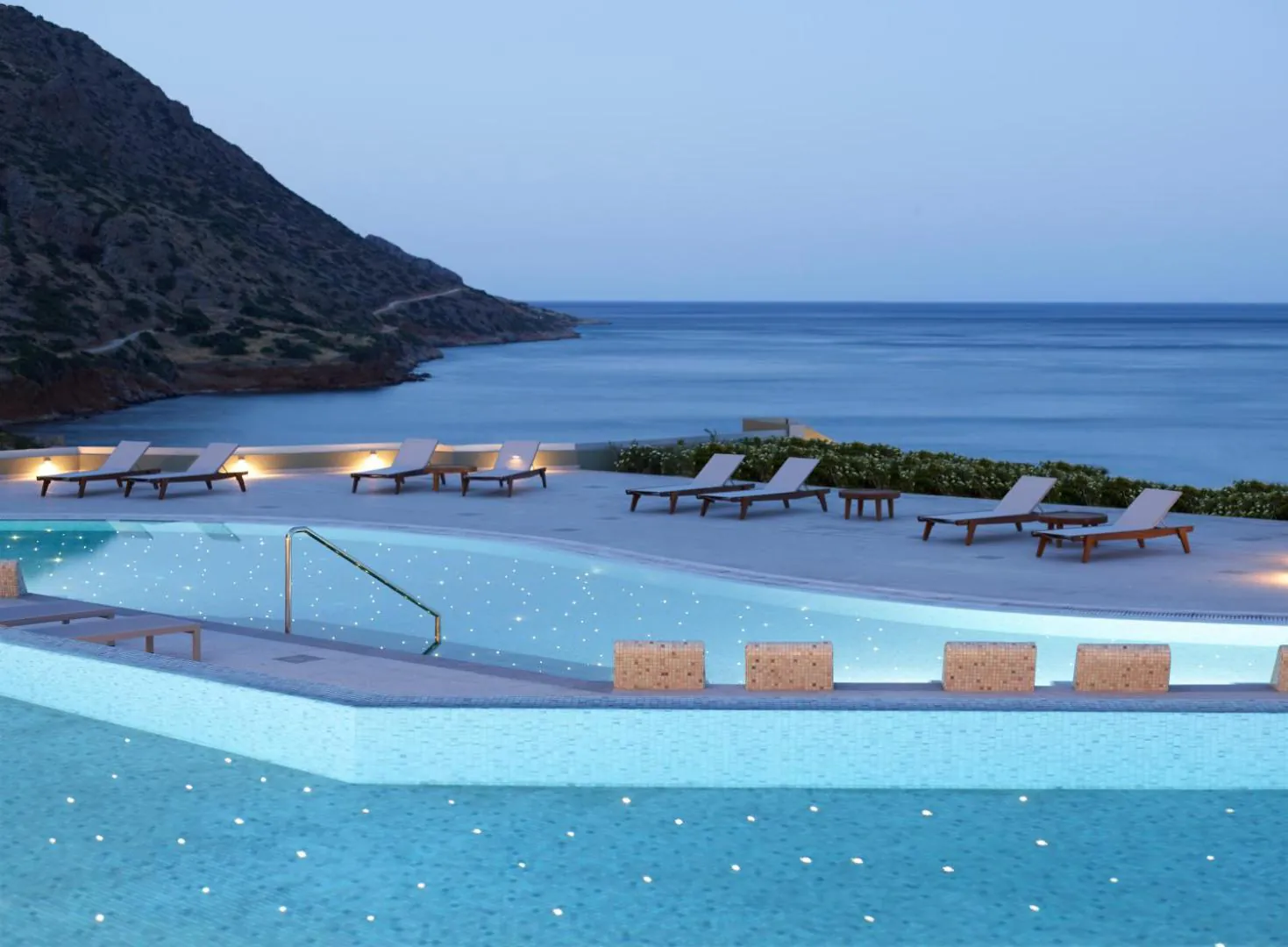 Infinity pool at dusk with loungers along the edge and the bay of Elounda below