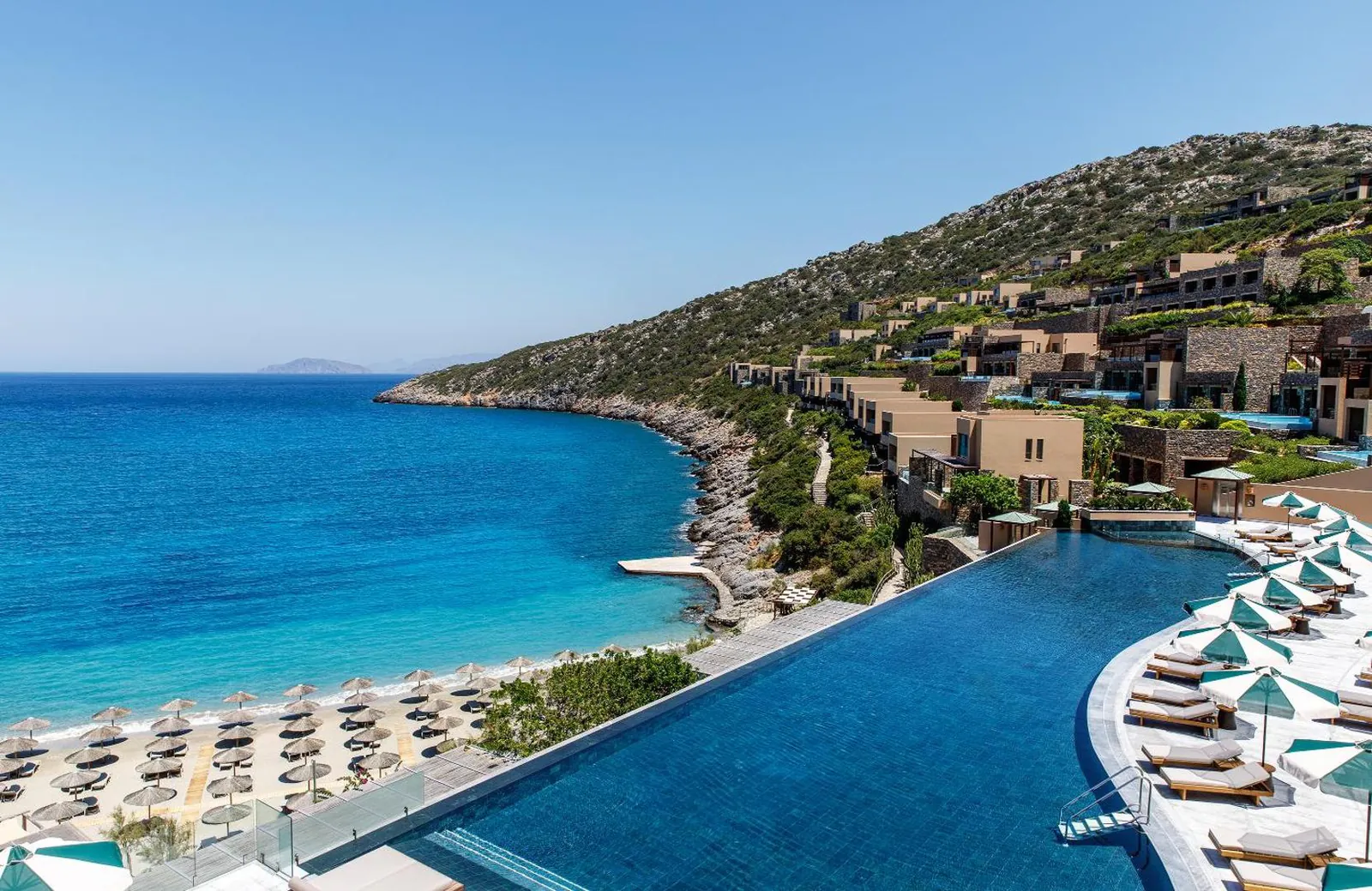 Infinity pool above Vathi cove at Daios Cove, with the sandy beach and the hillside resort behind