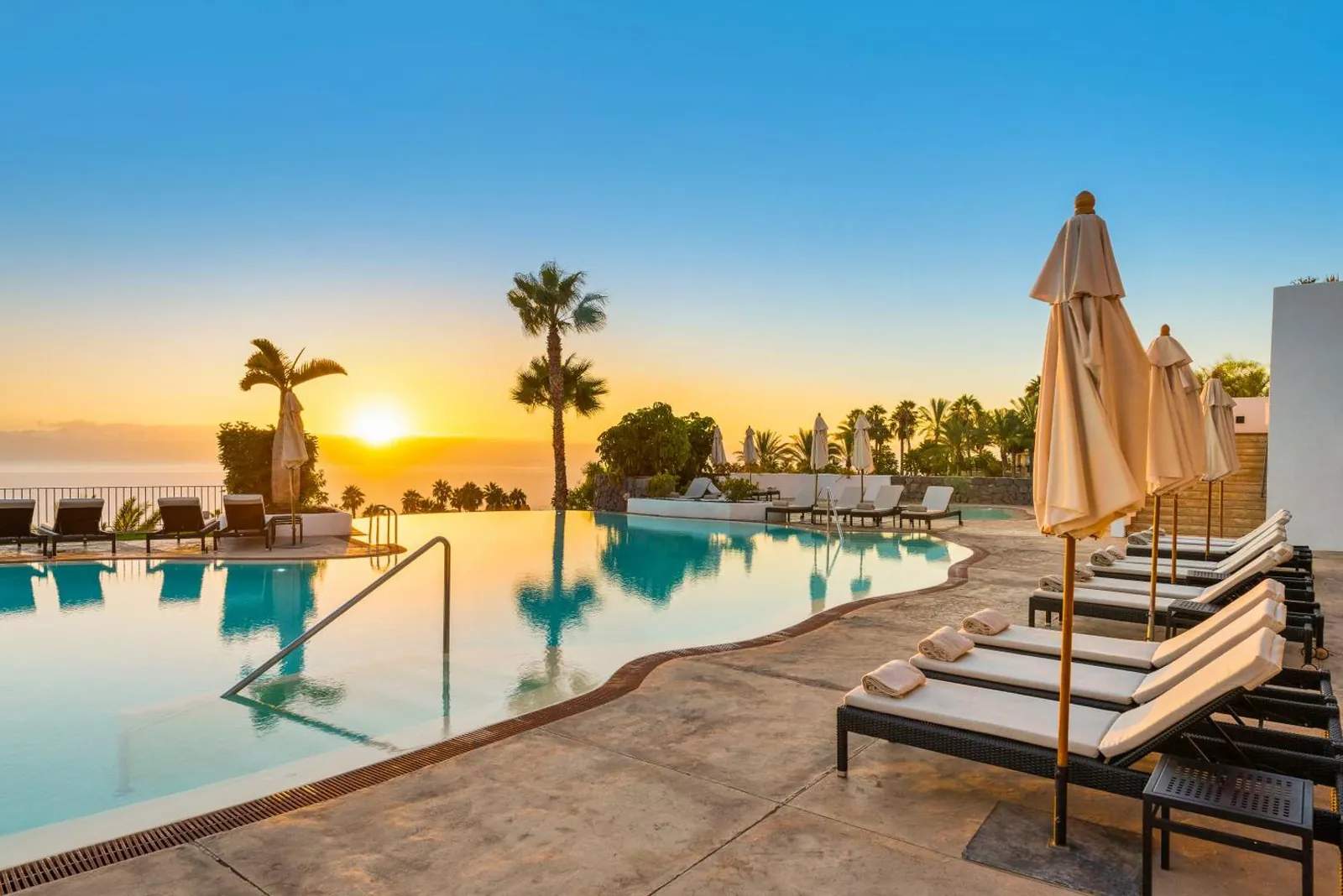 Infinity-edge pool at sunset with parasols and palms against the sea