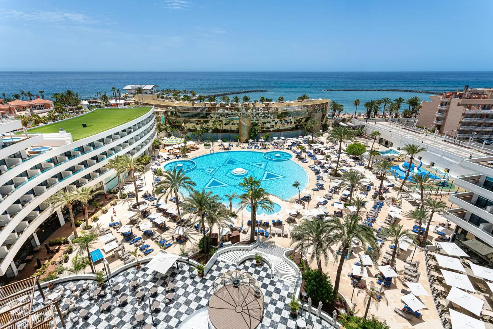 Lagoon-style pool among palms and loungers, curved hotel wings around it and the Atlantic beyond