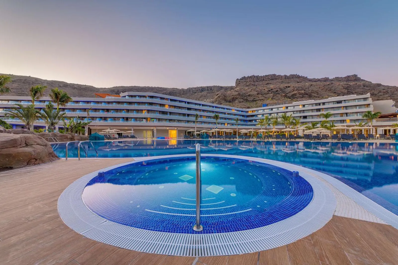 Round hot tub in front of the main pool at dusk, with the resort and Mogán's mountains behind