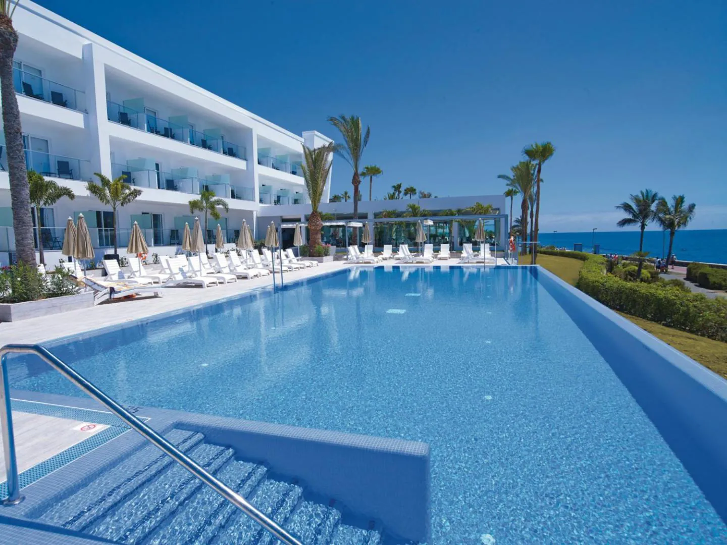 Long infinity pool beside the white hotel wing, with sun loungers along the deck