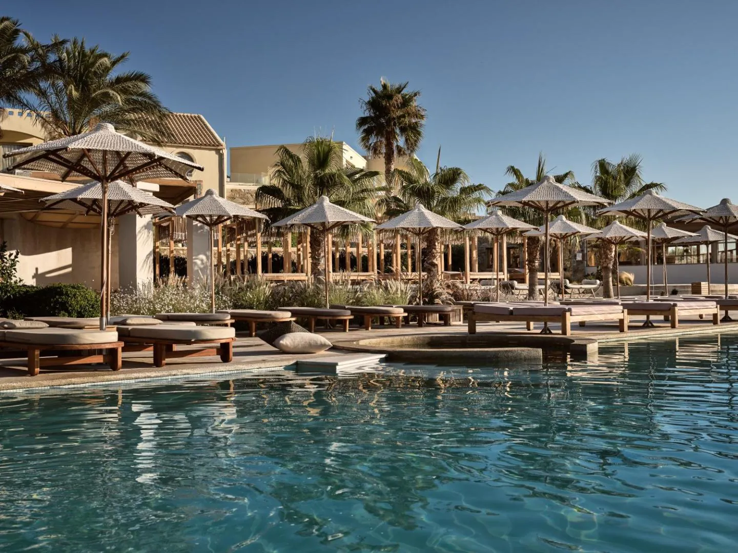 Outdoor pool edged with palms, wooden daybeds and thatched parasols